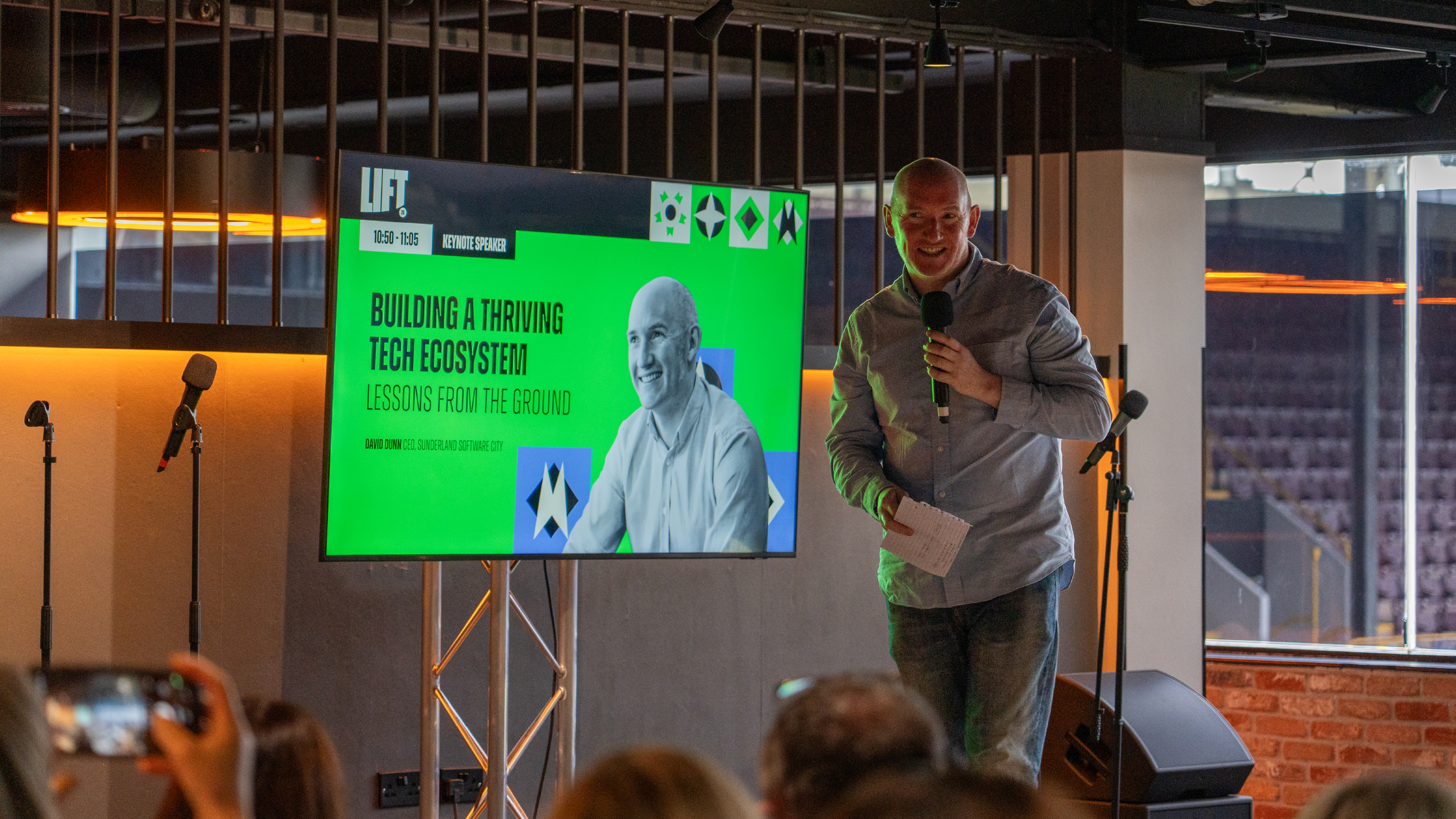 David Dunn on stage at LIFT26 - behind him are branded graphics on a screen introducing his keynote talk.