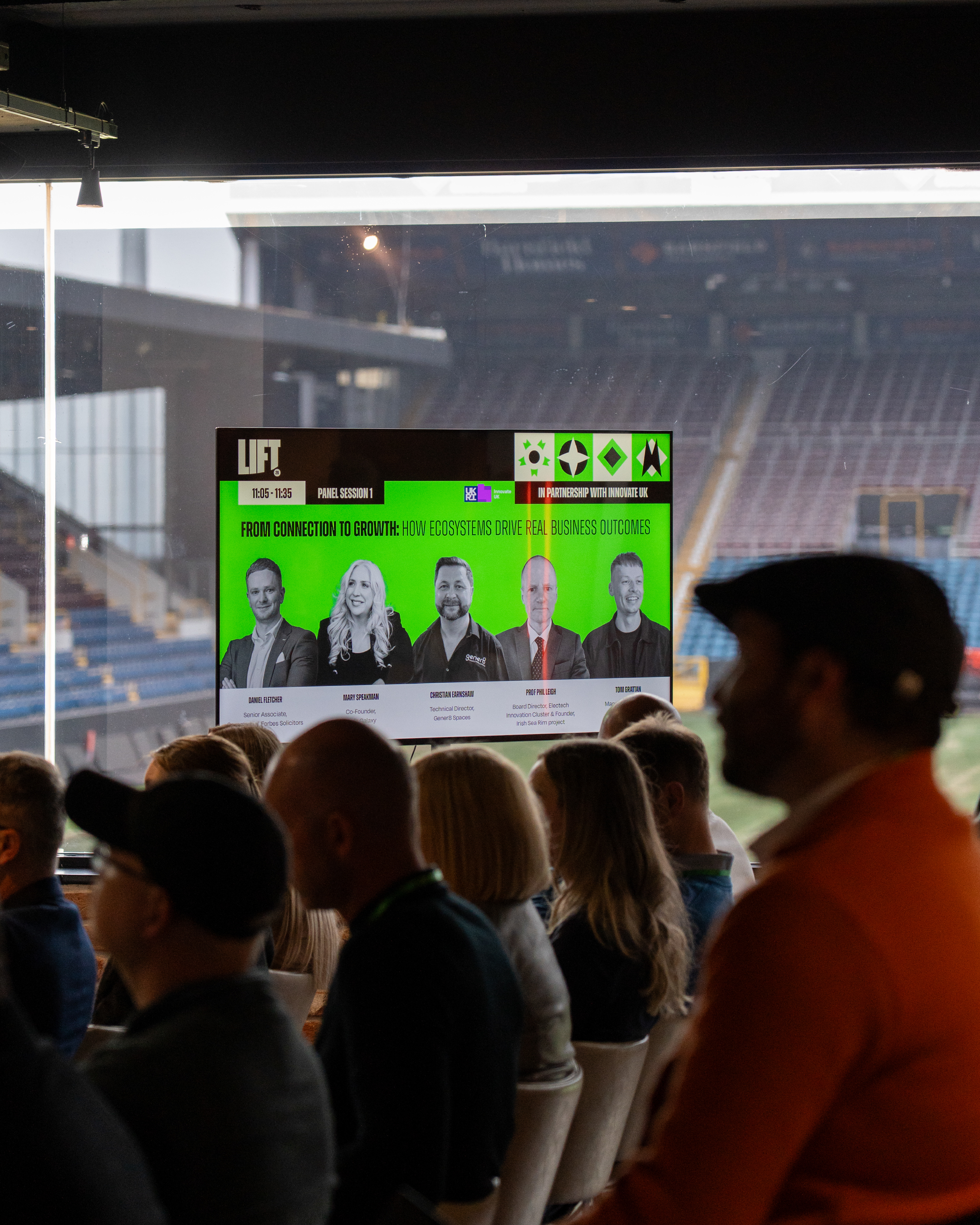 Panel session speakers displayed on a screen, in front of Turf Moor pitch and stands.