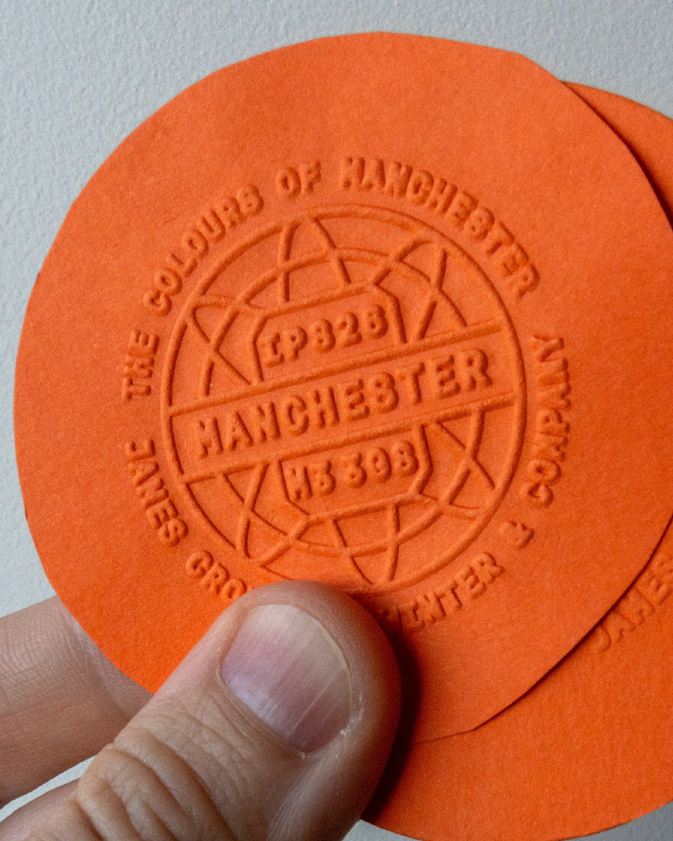 A close up of the emboss detail on Mandarin Orange paper. The design features 'The colours of Manchester' in a roundel design.